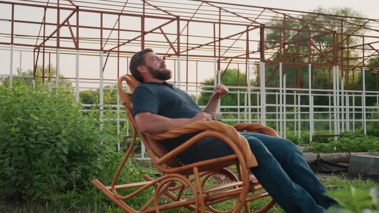 gardener seated on wooden rocker chair holding clear mug of herbal tea leaning back gazing into distant garden under warm sunset light surrounded by leafy plants metal trellis