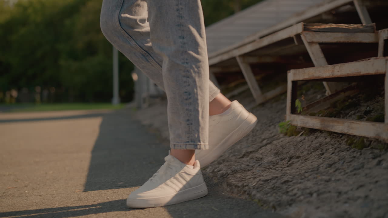 Close-up of woman s legs as she walks down rustic stadium seating, with white sneakers stepping on weathered metal and greenery sprouting among the rusty structure