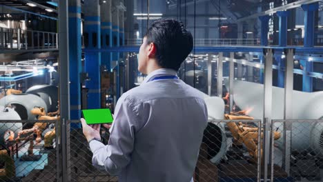 back view of an asian male professional worker standing at the center of the wind turbine factory, checking on smartphone with green screen and look at the factory once