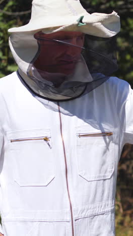 Beekeeper in protective suit outdoors. Man apiculturist working on apiary in summer day. Man in white uniform and hat near beehives on green nature background. Vertical video
