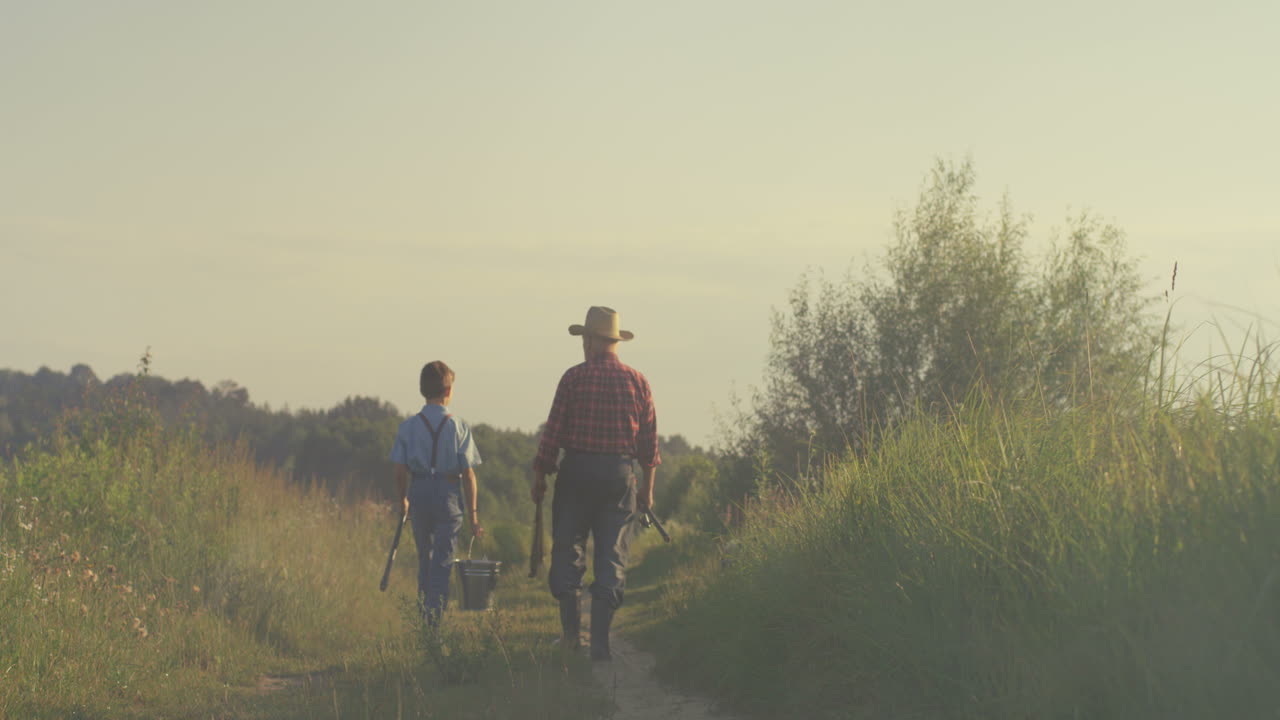 Rear view of caucasian grandfather and his little grandson walking together while holding fishing rods in the morning
