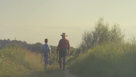 vista trasera del abuelo caucásico y su nieto caminando juntos mientras sostienen cañas de pescar por la mañana