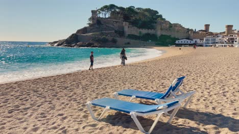 Liegestühle-Am-Strand-Mit-Dem-Hintergrund-Der-Ummauerten-Burg-über-Dem-Meer-In-Tossa-De-Mar,-Girona,-Spanien,-Costa-Brava,-Türkisfarbene-Wasserstrände