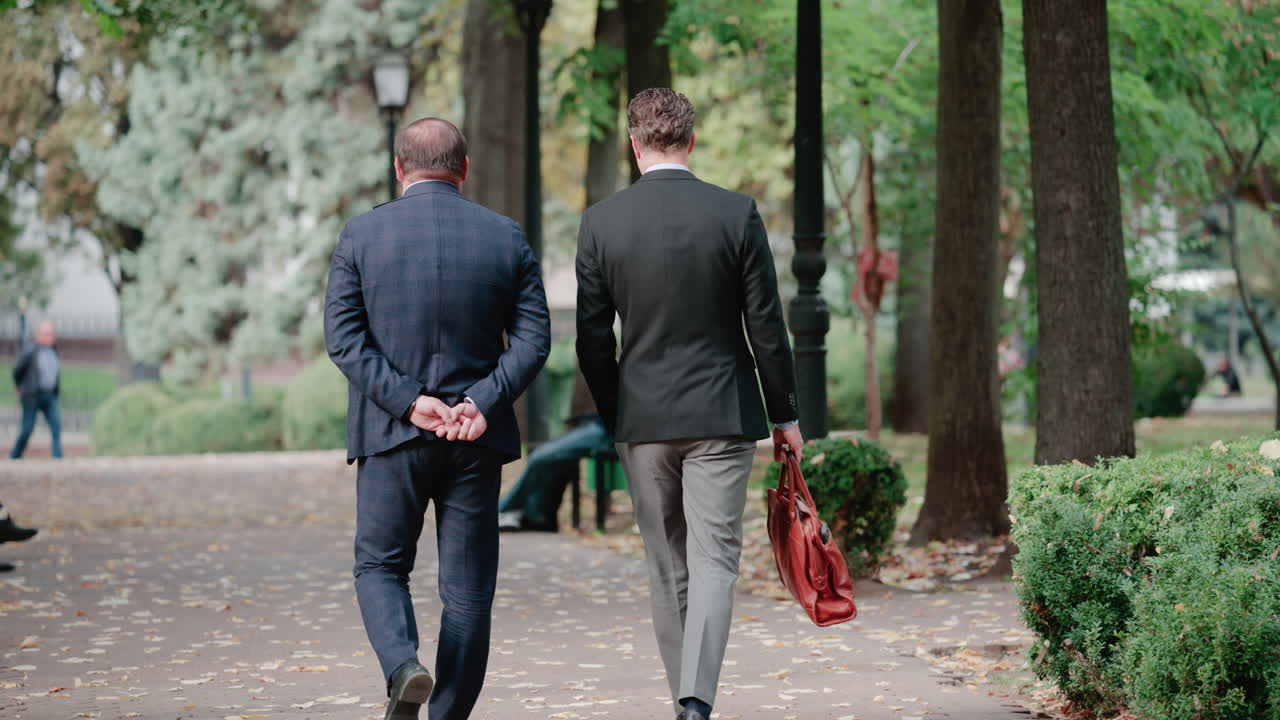 Two men in suits walk together through a leafy park, engaged in a business conversation during a calm autumn day