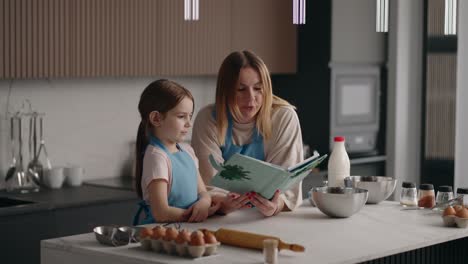 madre y hija caucásicas están cocinando en la cocina de casa el fin de semana, leyendo un libro de cocina