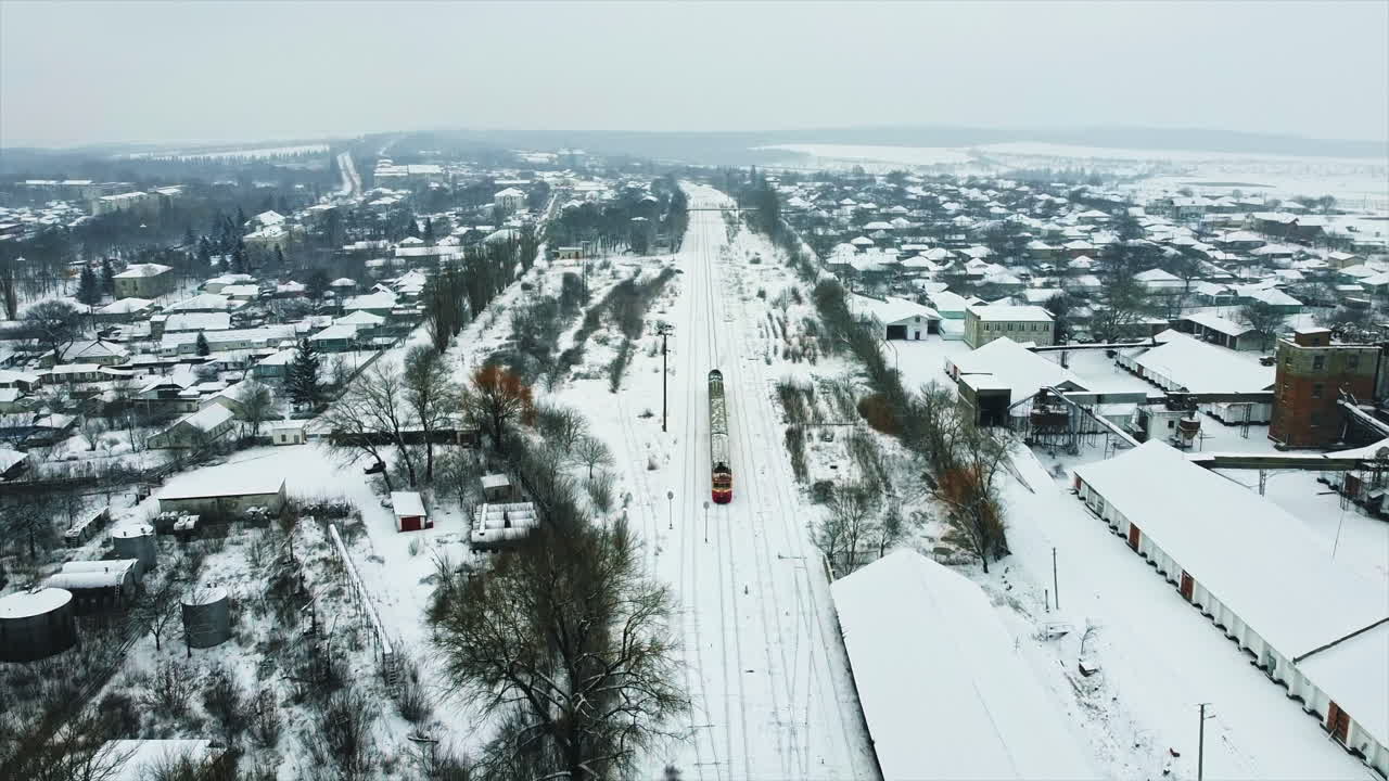 Aerial drone view of a moving train in a village, winter, Moldova. Railroad, station, residential buildings, bare trees, snow