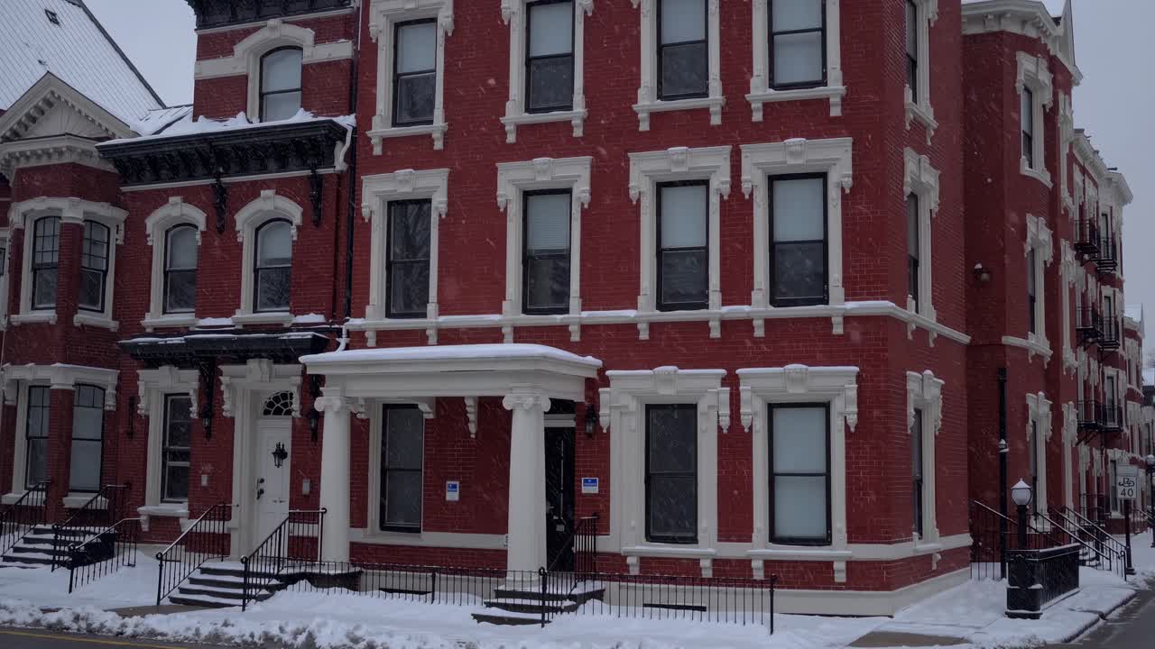 Snowflakes drifting over historic red brick residence, highlighting white architectural details, ornate windows, and classic porch during serene winter moment
