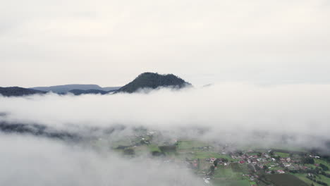 Vuelo-Aéreo-Con-Drones-A-Través-De-Las-Nubes-Con-Un-Pico-De-Montaña-En-El-Fondo-Con-Un-Pequeño-Pueblo-Debajo