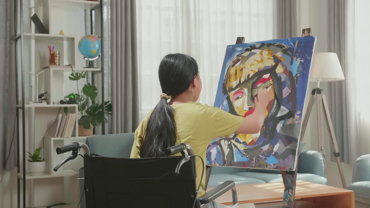 Hind View Of An Asian Artist Girl In Wheelchair Holding Paintbrush And Painting A Girl'S Face On The Canvas
