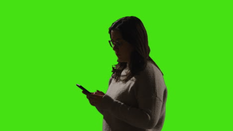 mujer entrando en el cuadro mirando un mensaje de texto o navegando en línea en el teléfono móvil contra un fondo de pantalla verde con iluminación de bajo nivel