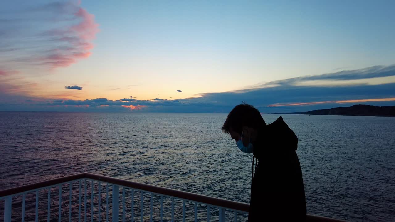 마스크와 재킷을 입은 관광객의 실루엣, 배경에 바다와 화려한 구름이 있는 매혹적인 풍경을 관광