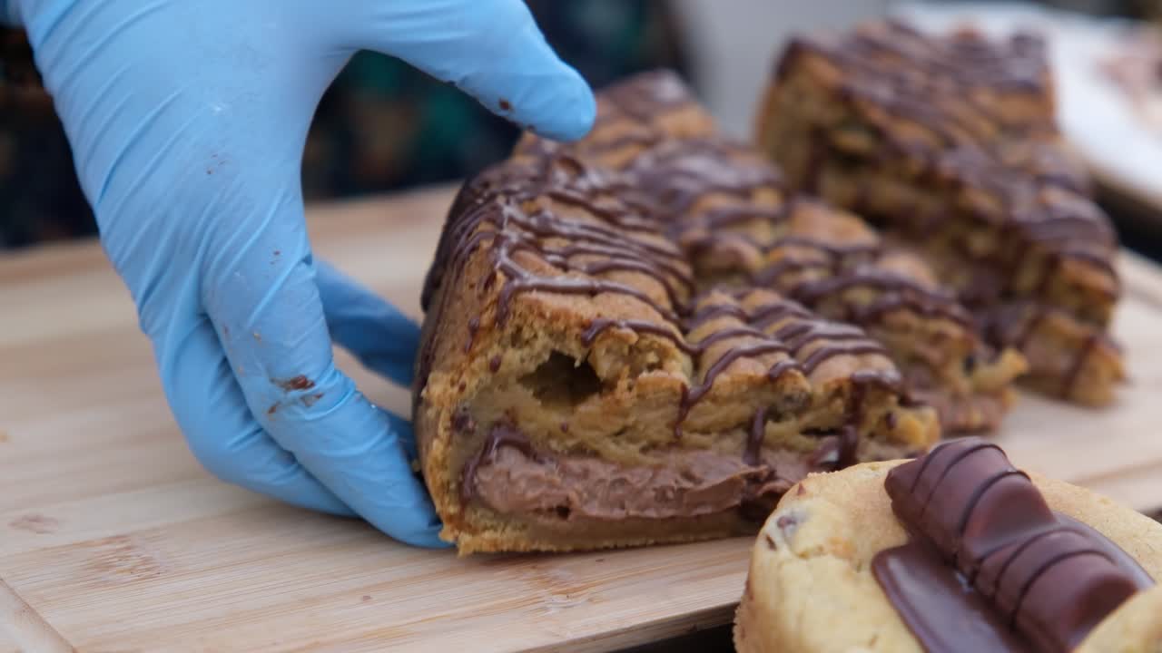 Close-up shot of a gloved hand lifting a thick cake slice filled with chocolate mousse from a wooden board. Features soft cake layers, rich mousse filling and chocolate drizzle in a bakery display