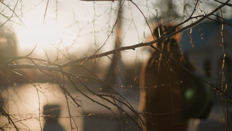 a sunny, blurred background frames a dry tree branch in the foreground, with a back shot of a woman wearing a brown coat, jeans, and white shoes, carrying a backpack as she walks through a park