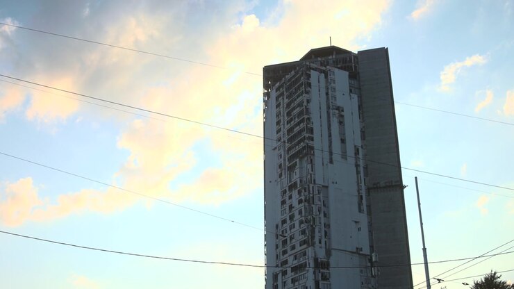 Um arranha-céu residencial alto e desgastado domina a vista da cidade, apresentando janelas empilhadas, grade de varandas, uma estrutura no telhado, antena, poço de serviço vertical e fios de utilidade diagonais, tudo sob um forte brilho suave e pastel do pôr do sol.
