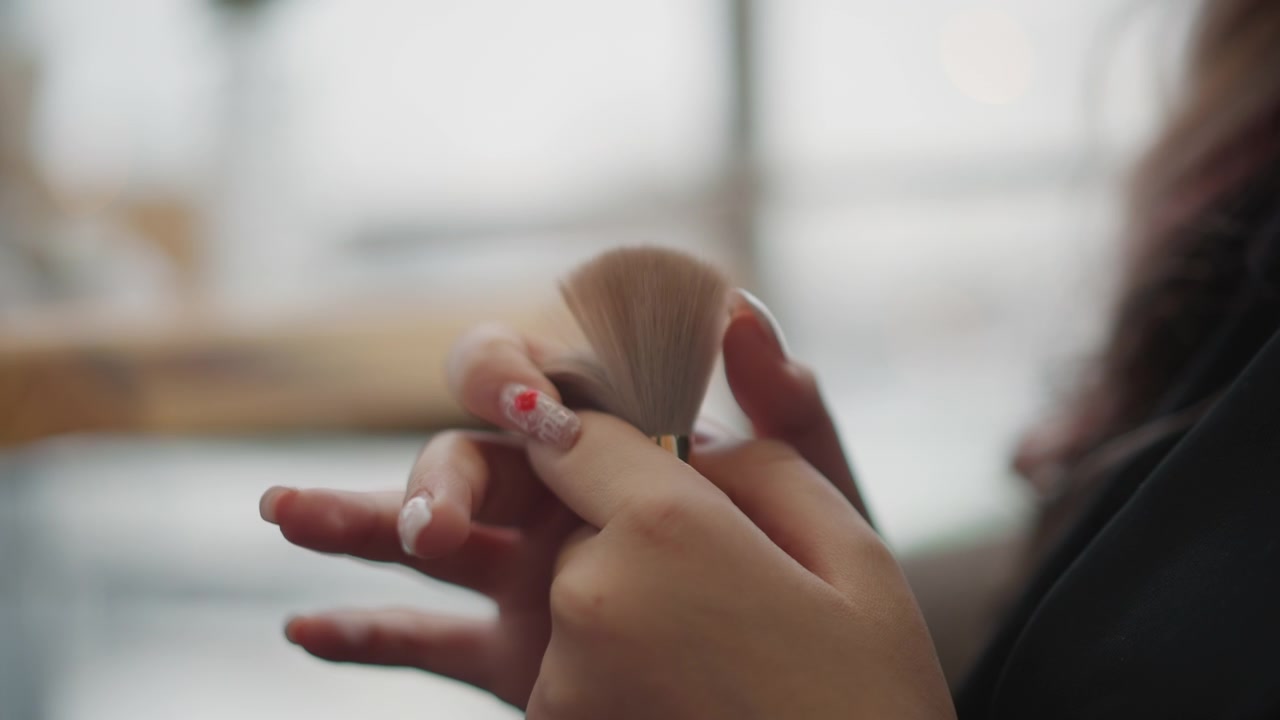 Close view of hands holding large fluffy makeup brush while sweeping bristles across fingertip, fingers slightly separated and thumb pinching base during careful inspection in soft window light