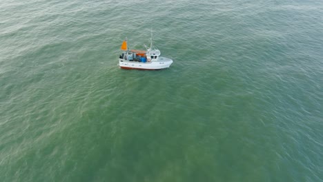 Aerial-view-of-a-coastal-fishing-boat-sailing-in-the-calm-Baltic-sea,-fisherman-dropping-fishing-net-in-the-water,-sunny-day,-distant-white-sand-beach,-wide-birdseye-drone-shot-orbiting-right