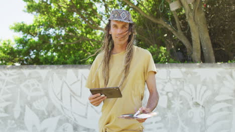 video shows cheerful caucasian man with dreadlocks, tablet, and palette by mural