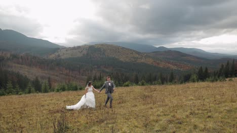 Lovely-young-newlyweds-bride-groom-running-on-mountain-slope,-holding-hands,-wedding-couple-family