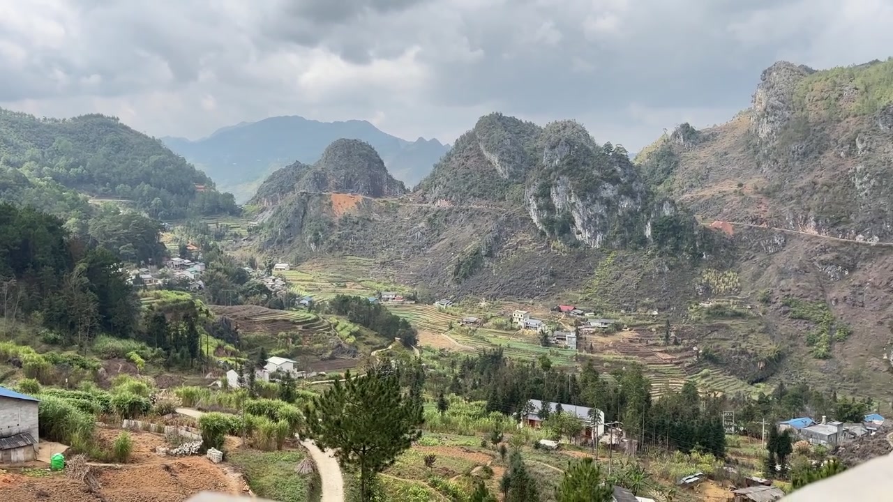 Pemandangan luas jalan pegunungan yang melewati tebing kapur dan lereng bukit bertingkat dengan tanah merah di pedesaan Ha Giang, Vietnam.