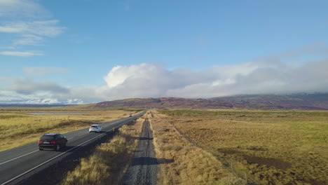 Drone-view-of-dirt-road-and-highway-with-vehicles-next-to-fields-and-farmland-with-mountains-in-the-background