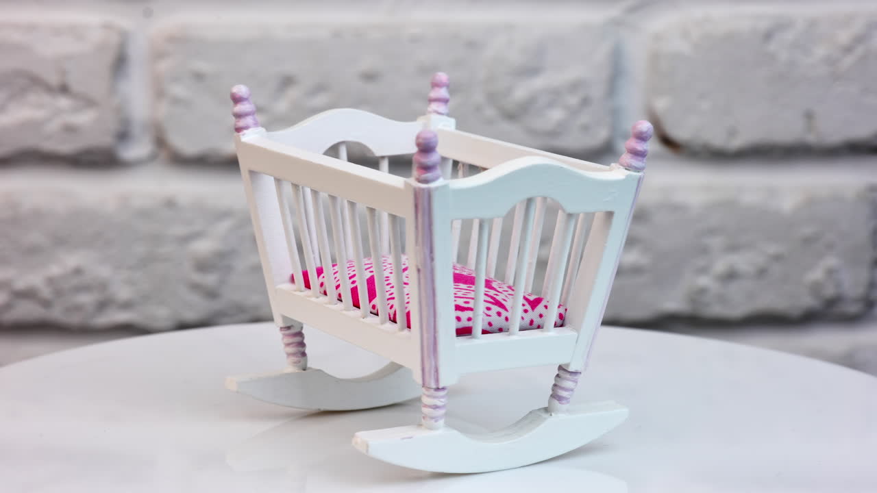 Beautiful toy cradle on a turning table. White baby crib made of wood with pink mattress as a piece of toy furniture. White brick wall at the backdrop.