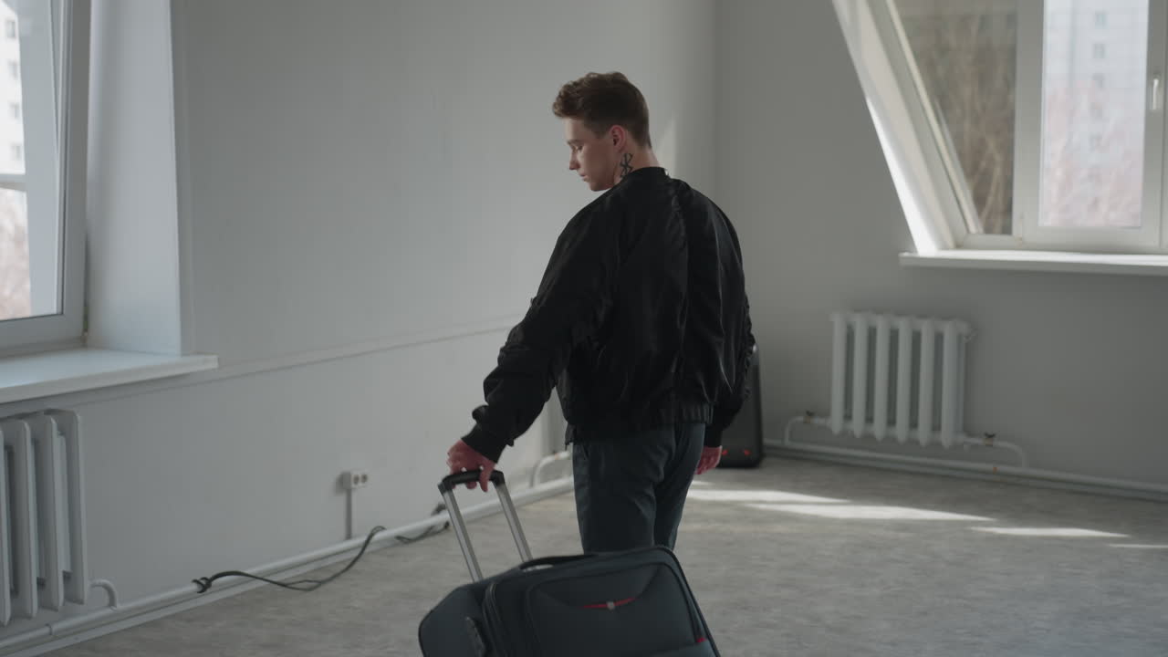 Man with neck tattoo pushes large wheeled travel bag to front while standing in bright minimalist room with radiator and window in background