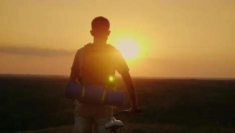 un hombre con una mochila y una bicicleta. se para y mira al horizonte donde se pone el sol. ciclismo y aventura