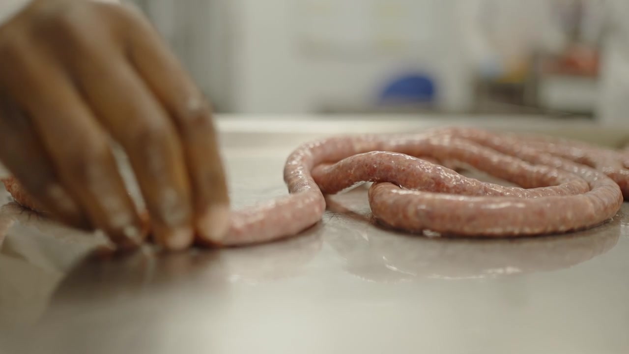 A continuous raw sausage strand is guided and arranged by hand across a wet metal table in a meat processing plant.