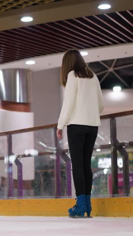 primer plano de una mujer con patines azules, pantalones negros y una camiseta blanca mientras patina en la pista de patinaje, con otras personas patinando