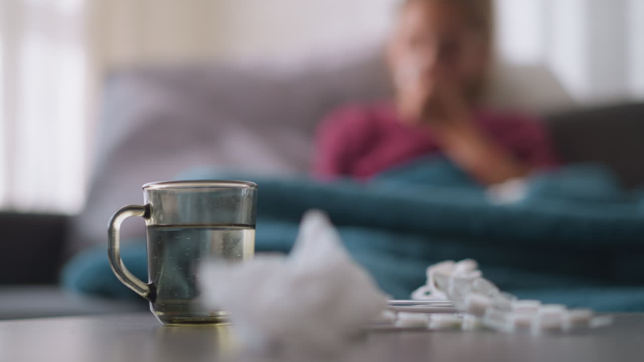 Close-up of medication, used tissue, and glass cup on table, with blurred view of sick person in background resting, showing signs of cold or flu symptoms, looking unwell