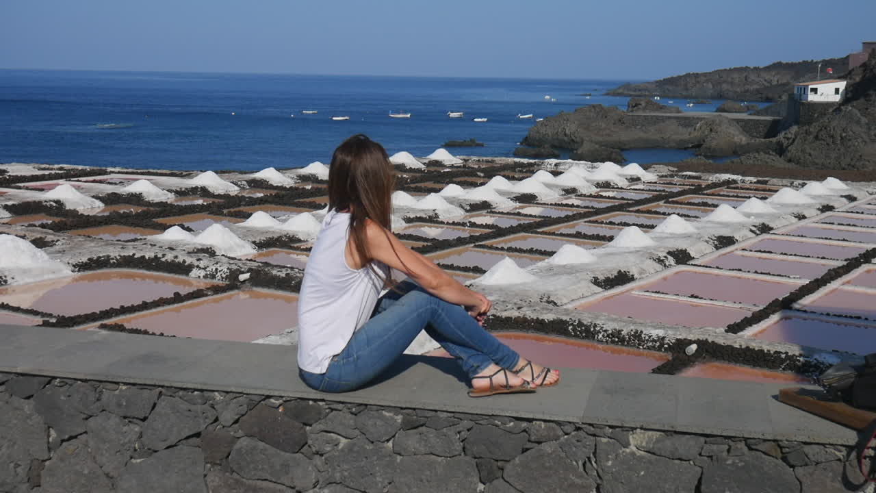 Caucasian woman sitting on a stone wall watching the salt pans and the Atlantic Ocean in La Palma