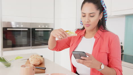 Multitasking in kitchen, asian woman eating sandwich and using smartphone, at home