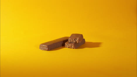 hand lifts one of several stacked vegan protein bars with a chocolate coating and visible soft, dense interior. yellow background, ingredients like soy, pea, rice, and milk protein fitness-focused.