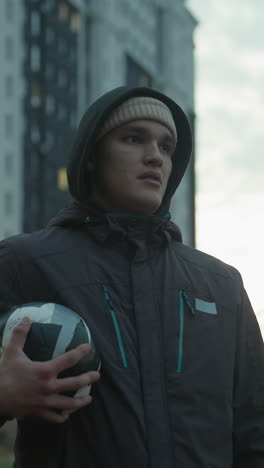 atleta sosteniendo una pelota de fútbol respira profundamente durante una sesión de entrenamiento urbano, de pie con confianza en una moderna arena deportiva rodeada de altos edificios residenciales y un espectacular cielo nocturno