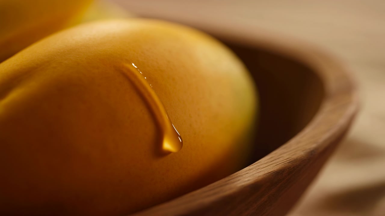 Primo piano macro estremo di mango giallo maturo all'interno di una ciotola di legno poco profonda, che mostra una singola gocciolina lucida che scivola lentamente lungo la buccia curva con luce calda, texture fine, ombra morbida e ricca naturalezza