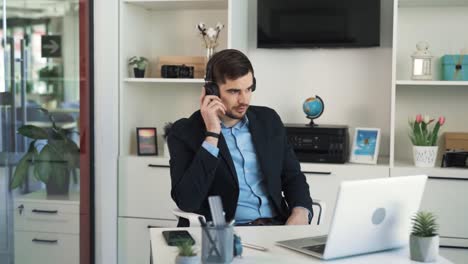 The-young-attractive-man-is-listening-to-a-new-song-in-his-headphones-in-a-bright-office