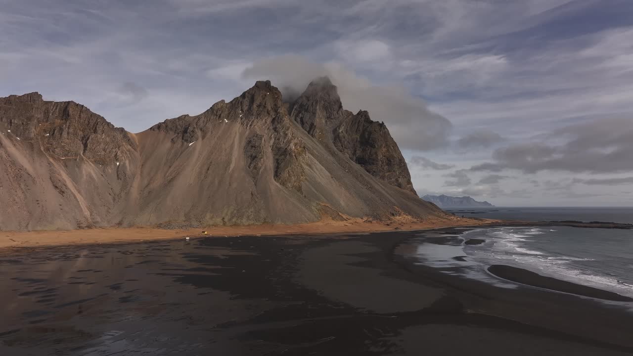 Aerial view of Vestrahorn mountain range and black sand beach with soft waves and cloudy sky, Stokksnes, Iceland.