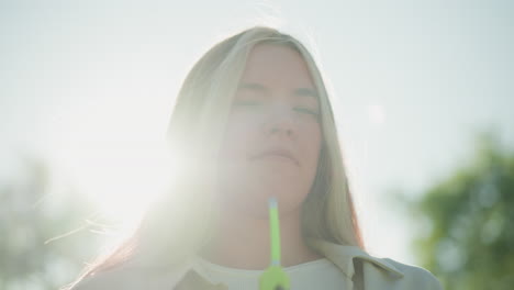 close-up of young woman standing in bright sunlight, holding green air pump to cool her face, with her blonde hair gently fluttering in the breeze, her expression is calm and focused