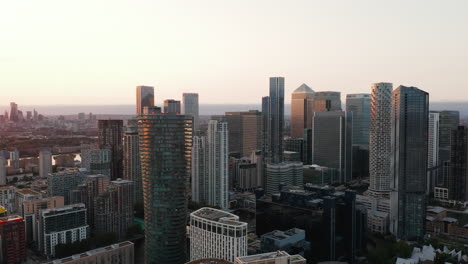 Descending-aerial-view-of-group-of-skyscrapers-in-Canary-Wharf-financial-and-economic-hub.-Modern-city-district-lit-by-setting-sun.-London,-UK