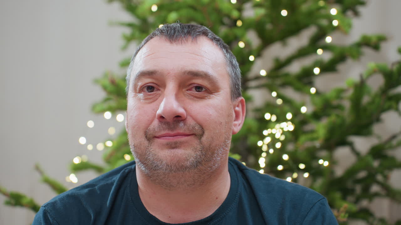 Portrait view of cheerful middle aged man in green top smiling warmly with decorated christmas tree glowing in background capturing cozy festive atmosphere during joyful family holiday season indoors