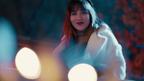 hermosa mujer de cabello oscuro de pie detrás de la valla sonriendo, rodeada de luces de navidad decoración durante una fría noche de invierno