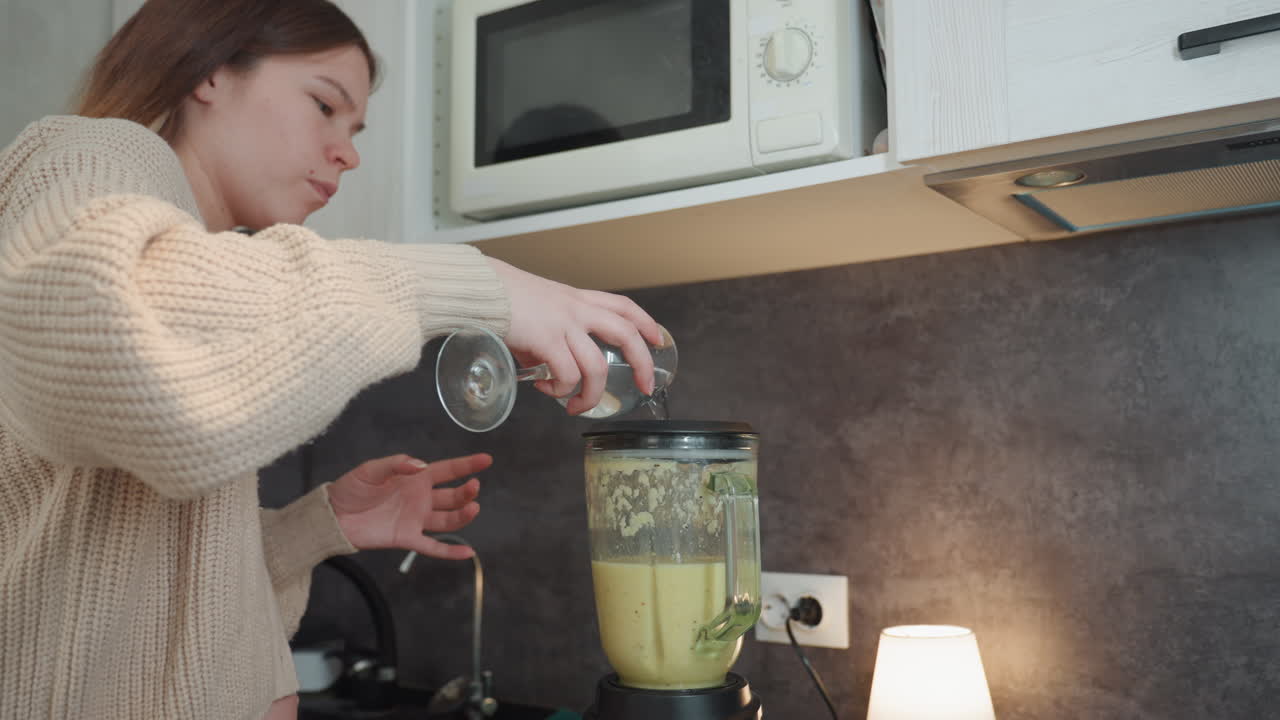 Donna bianca incinta versa ingredienti nel frullatore, indossando un maglione accogliente in cucina. Il pancione è visibile, una lampada sul bancone illumina la scena. Con misurazioni accurate e calma concentrazione, prepara un frullato prenatale per la casa.