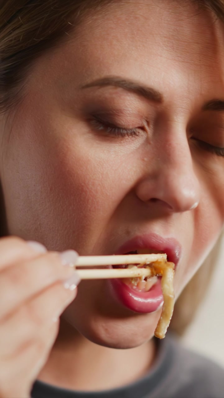 Close up of woman using chopsticks to eat noodles. subject hand and wooden utensils are visible as woman brings food to her mouth with soft lighting highlighting skin texture and subtle