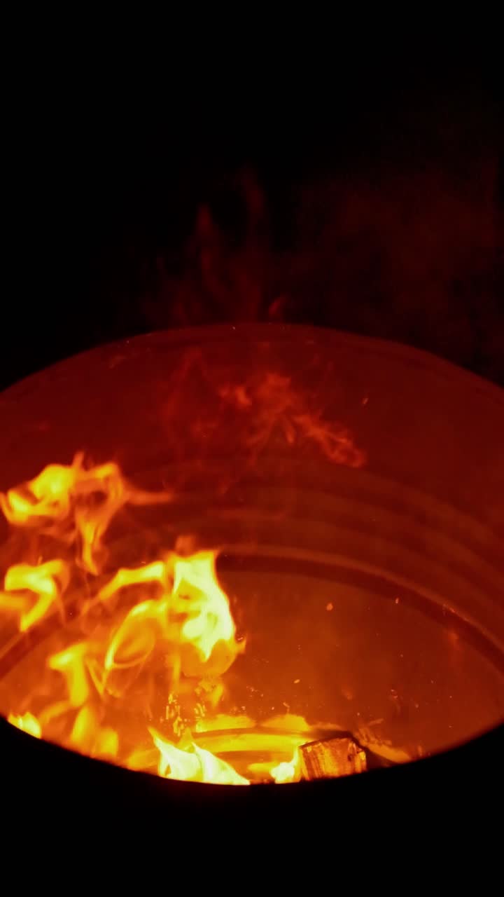 Close-up cinematográfico 4K da mão de uma pessoa colocando um pedaço de madeira em uma fogueira de acampamento crepitante à noite.