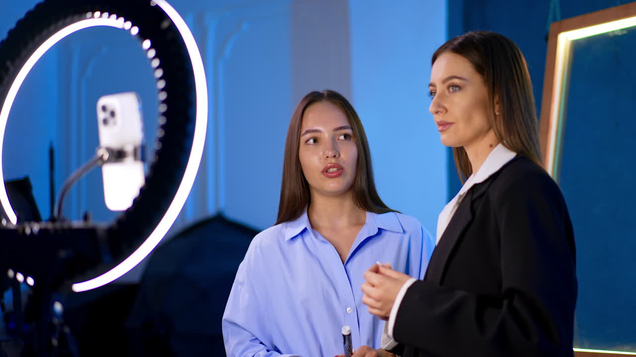 Chatty beauty tutorial. Two women converse in a studio setting, preparing for a beauty tutorial with ring light and smartphone setup