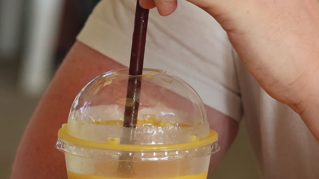 A close-up shot of a person's hand stirring a yellow smoothie in a plastic cup