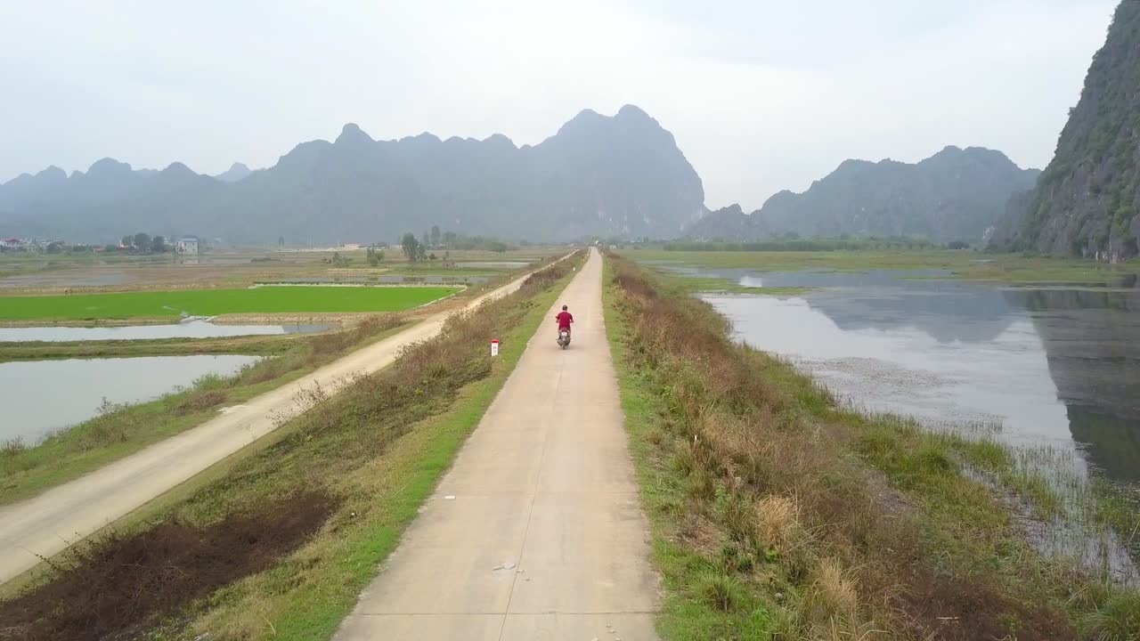 Drone follow cam of motor biker in red driving on narrow road surrounded by grass, rice fields and reflecting lake with tall mountains in background