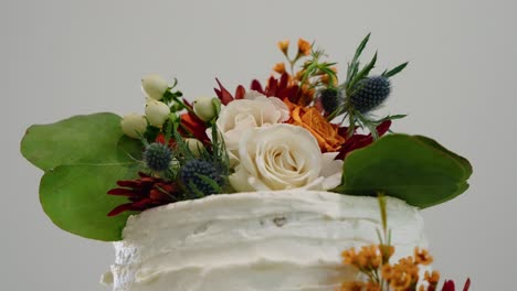 a detailed shot of a floral wedding cake topper featuring white roses, orange flowers, and blue thistles. the rustic-style cake showcases elegant floral decor.