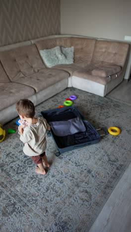 small kid picks clothes from the floor scattered around the suitcase. baby boy throws his things up with a joy. top view. vertical video.