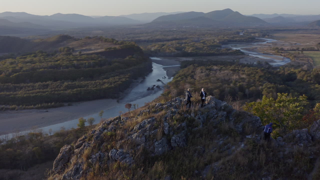Aerial shot of  people standing  on top of a huge cliff with the view on valley with riverbed taking a one hundred eighty degree turn and mountain ridge  in the background on the sunset.